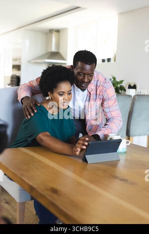 Penché sur l'épaule couple afro-américain à l'aide de tablette à la table de cuisine, avec des tasses à café. Technologie, connexion, style de vie, contemporain, domest Banque D'Images