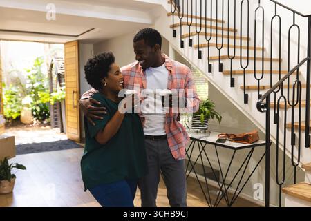 Couple afro-américain debout tenant des tasses de café à emporter dans le foyer, montrant sac à main et plante. Romantique, confortable, style de vie, moderne, domestique, connecti Banque D'Images