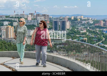 Deux femmes marchant sur un point de vue avec un paysage urbain panoramique de Vladivostok, Russie, des bâtiments résidentiels modernes et des collines verdoyantes en arrière-plan. Banque D'Images