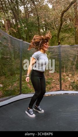 Une jeune femme heureuse vêtue de vêtements de sport avec ses jambes pliées et ses cheveux en l'air après avoir sauté sur un trampoline en plein air dans les montagnes, profitant Banque D'Images