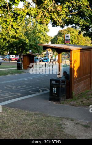 Une personne assise dans un abri de bus en bois à un arrêt de bus à Saffron Walden, Essex, Angleterre sous une canopée d'arbres. Banque D'Images