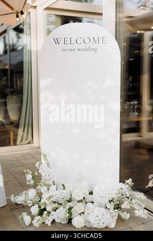 Un panneau de bienvenue magnifiquement conçu accueille les invités à un mariage, entouré d'élégantes fleurs blanches, créant une atmosphère romantique. Banque D'Images