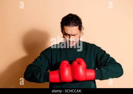 Jeune homme en Vert Pullover portant des gants de boxe sur fond beige exprimant des émotions confiantes et déterminées Banque D'Images
