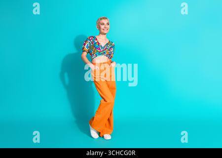 Jeune femme élégante avec des cheveux blonds courts portant une tenue boho colorée sur fond turquoise vibrant souriant joyeusement Banque D'Images