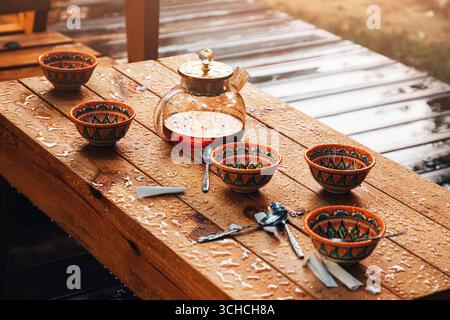 Théière en verre et tasses décorées sur une table en bois humide sous la pluie, créant une atmosphère chaleureuse et rafraîchissante Banque D'Images