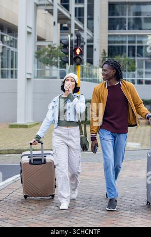 Couple diversifié marchant à travers la rue tirant des bagages et sirotant de tasse de café en traversant le signal. Urbain, voyage, moderne, style de vie, décontracté, transpo Banque D'Images