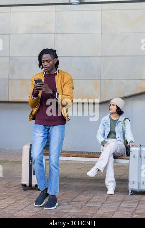 Couple diversifié debout sur la plate-forme de la station avec des valises et vérification smartphone, espace de copie. Urbain, transport, voyage, moderne, style de vie, conn Banque D'Images