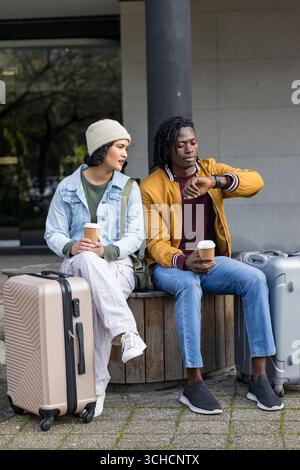 Couple diversifié assis sur le banc à l'entrée tenant des tasses à café vérifiant smartwatch près des valises. Romantique, loisir, urbain, moderne, style de vie, casua Banque D'Images
