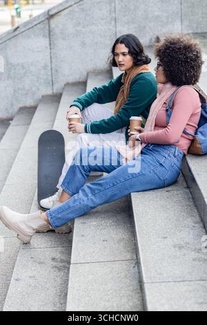 Des amies assis sur les marches urbaines portant des foulards et sirotant un café à emporter près de skateboard. Occasionnel, amitié, urbain, style de vie, moderne, vibra Banque D'Images