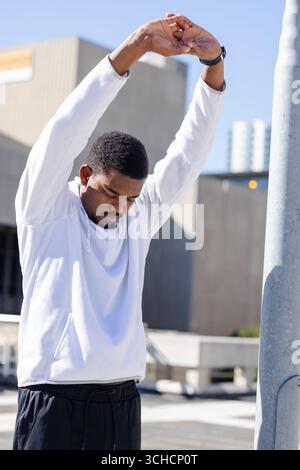 Homme afro-américain étirant les bras au-dessus de la tête sur le toit en pantalon noir à capuche blanc et montre-bracelet. Urbain, fitness, style de vie, moderne, actif, outdoo Banque D'Images