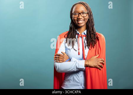 Joyeux super-héros féminin noir se tient avec les bras croisés, regardant la caméra sur fond isolé. Femme afro-américaine souriante portant une chemise bleue et une cape rouge, posant en toute confiance en studio. Banque D'Images