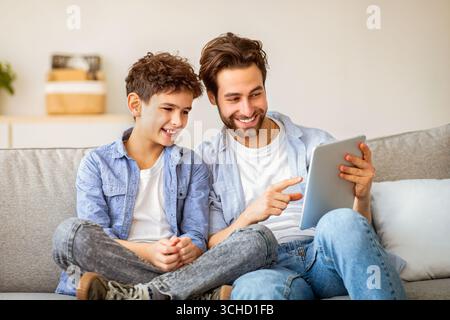 Père et fils joyeux utilisant une tablette, regardant des vidéos ou jouant à des jeux en ligne, passant du temps ensemble à la maison, Banque D'Images