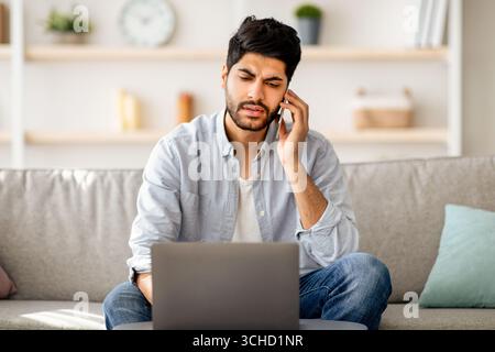 Activités à distance. Inquiet indépendant arabe parlant sur téléphone portable et utilisant ordinateur portable, assis sur un canapé à la maison Banque D'Images