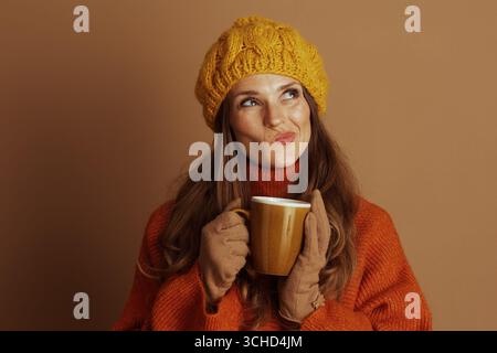 Une femme en béret tricoté jaune et col roulé orange tient une tasse, regardant vers le haut avec une expression réfléchie. La scène transmet aut confortable et réfléchissant Banque D'Images