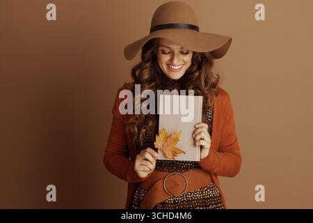 Une femme élégante à la mode automnale, portant un cardigan rouille et un chapeau à larges bords, sourit tout en tenant un livre rose orné d'une feuille jaune d'automne aga Banque D'Images