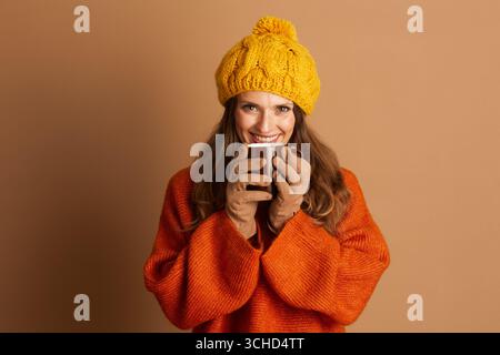 Une jeune femme adulte souriante avec de longs cheveux bruns, en pull orange tricoté et bonnet jaune pompon, tient une tasse brune chaude près de sa bouche. Elle regarde Banque D'Images
