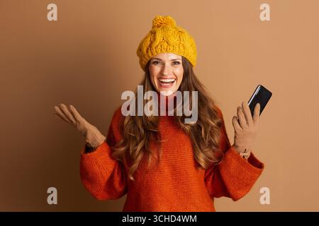 Une femme joyeuse portant un chapeau à pompon et un pull à col roulé orange sourit avec éclat, tenant son smartphone et faisant des gestes excités. Elle transmet l'enthousiasme et Banque D'Images