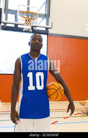 Homme afro-américain en maillot bleu tenant le basket-ball sous le bras droit sur le terrain de gymnastique avec cerceau Banque D'Images
