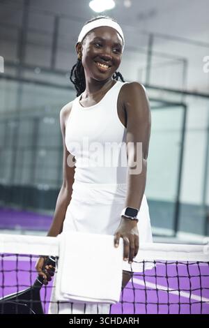 Femme afro-américaine debout au filet de tennis sur un court couvert sous les lumières, tenant la raquette Banque D'Images