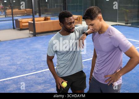 Divers amis masculins s'appuyant sur des raquettes, tenant le ballon, discutant sur le terrain de padel près des canapés en cuir Banque D'Images