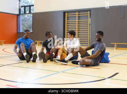 Divers amis masculins assis sur le sol de la salle de gym tenant basket-ball et bouteille d'eau à côté des bancs Banque D'Images