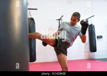 Homme afro-américain kicking sac de punching dans le studio de boxe sur des tapis roses dans des gants de boxe Banque D'Images