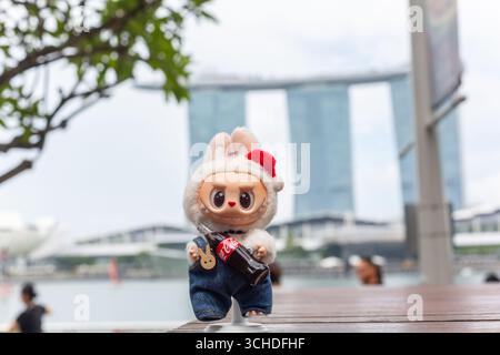 Figurine Labubu de couleur blanche tenant une bouteille de coke posant contre l'hôtel Marina Bay Sands, emblématique et emblématique à visiter à Singapour. Banque D'Images