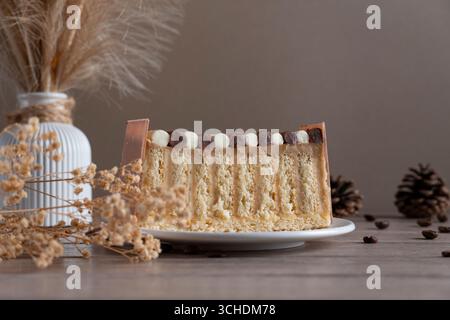 Un gâteau superposé magnifiquement tranché sur une assiette blanche, entouré d'un décor rustique comprenant des fleurs séchées, un vase blanc et des pommes de pin, créant un a confortable Banque D'Images