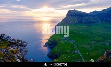 Des teintes orange et rose époustouflantes remplissent le ciel alors que le soleil se couche sur les îles Féroé. Cascade Mulafossur près du village de Gasadalur Banque D'Images