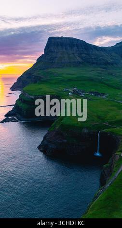 Vue imprenable sur la cascade de Mulafossur, qui plonge de façon spectaculaire dans l'océan Atlantique près du village de Gasadalur. Le coucher de soleil éclatant jette une lueur chaude sur le paysage accidenté. Banque D'Images