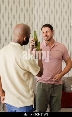 Divers amis masculins clinking bouteilles en verre vert sur le canapé du salon avec coussin rouge, espace de copie Banque D'Images
