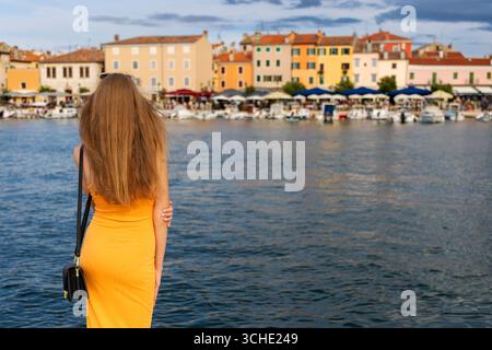 Femme en robe jaune admirant la vue sur la ville côtière. Jeune femme aux cheveux longs dans une robe jaune se tient près du front de mer, regardant les bâtiments colorés Banque D'Images