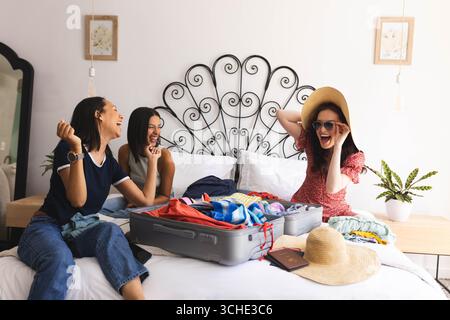 Diverses amies féminines riant et emballant la valise sur le lit, se préparant pour les vacances, à la maison Banque D'Images
