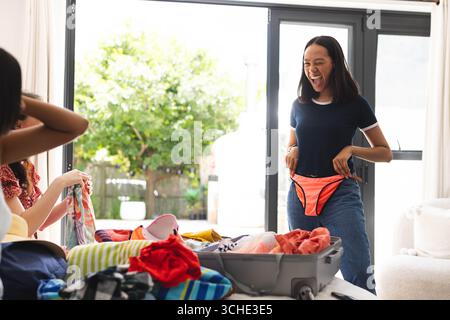 Diverses amies féminines emballant joyeusement des vêtements pour des vacances à la maison, choisissant avec enthousiasme des tenues Banque D'Images
