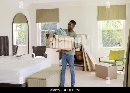 Portant homme afro-américain tenant une boîte en carton de vêtements dans la chambre, avec miroir et pouf Banque D'Images