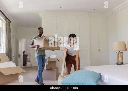Couple diversifié déballer des boîtes en carton dans une chambre lumineuse, avec des œuvres d'art encadrées et des couvertures Banque D'Images