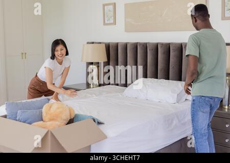 Faire le lit diversifié couple lissant les draps blancs dans la chambre, avec ours en peluche et oreillers Banque D'Images