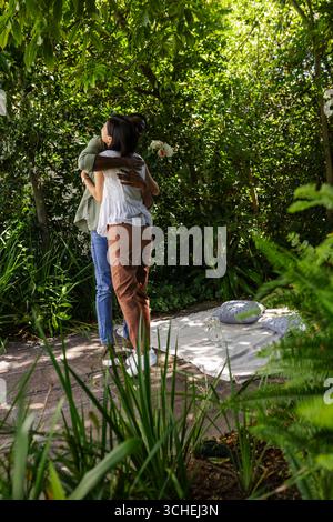 Embrassant un couple diversifié debout sur un chemin de pierre dans un jardin verdoyant, avec une couverture de pique-nique et des fleurs pâles Banque D'Images