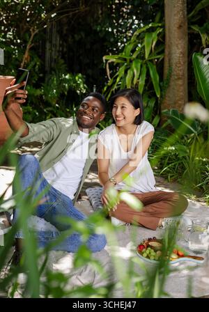 Prise selfie couple diversifié assis sur une couverture de pique-nique dans le jardin de l'arrière-cour, avec smartphone Banque D'Images