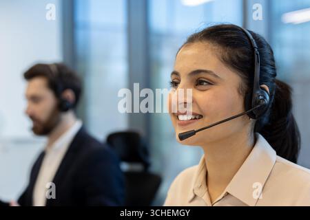 Représentante du service client souriant et parlant avec un micro-casque tout en travaillant dans un centre d'appels occupé, fournissant assistance et assistance aux clients Banque D'Images