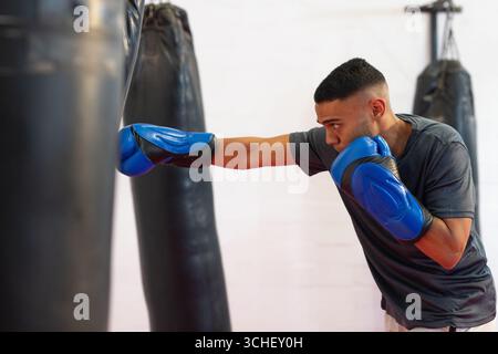 Homme poinçonnant sac lourd en chemise grise et gants de boxe bleus dans la salle de boxe, espace copie Banque D'Images
