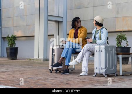 Divers amis assis sur un banc en bois à la place extérieure avec valises à roulettes, smartphone Banque D'Images
