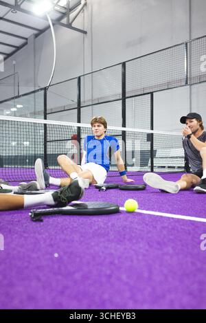 Amis masculins assis sur un court de padel violet derrière un filet bas reposant avec des raquettes, balle de tennis Banque D'Images