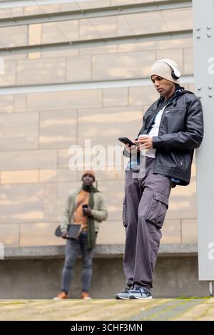 Divers amis masculins debout dans plaza vérifiant le téléphone avec des écouteurs et skateboard Banque D'Images