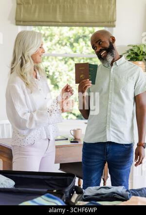 Souriant couple senior diversifié planifier voyage dans le bureau à domicile, tenant des passeports et une valise ouverte Banque D'Images