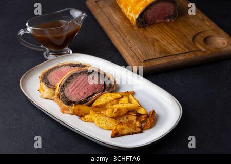 Wellington de bœuf avec tranche de pomme de terre cuite au four et sauce demi-glace. Plat festif traditionnel de filet de bœuf avec champignons et jambon de Parme cuit en feuilleté p Banque D'Images