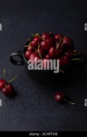Cerises fraîches mûres juteuses avec des gouttes d'eau dans une plaque noire sur un fond noir. Fruits de saison, récolte. Mise au point sélective, gros plan. Banque D'Images