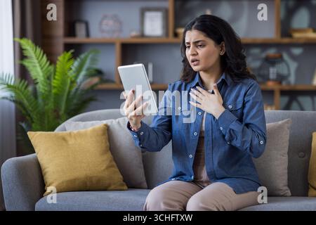 Inquiet jeune femme indienne debout sur le canapé à la maison et parlant en ligne via tablette. Banque D'Images