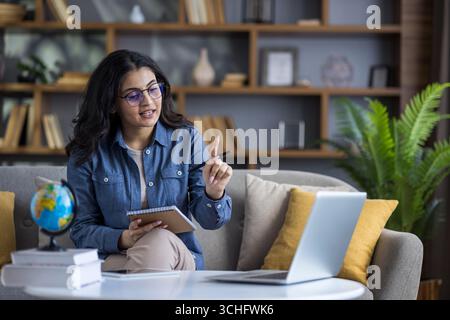 Jeune étudiante indienne assise sur le canapé à la maison, tenant un cahier et menant un apprentissage à distance via un ordinateur portable. Banque D'Images