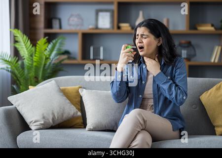 Jeune femme indienne assise sur un canapé à la maison, tenant son cou avec sa main, ressentant une douleur intense, en utilisant un spray médical. Banque D'Images